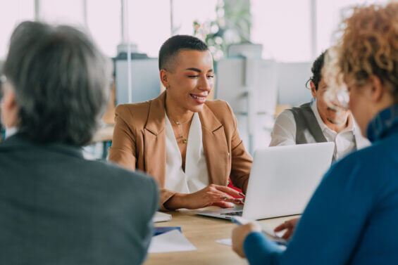 Portrait of smiling young businesswoman sitting in boardroom with her team and typing on a laptop during the meeting. Young experienced data analyst using laptop on a business meeting.