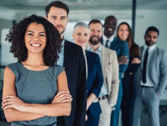 Portrait of beautiful smiling businesswoman with her colleagues. Multi-ethnic group of business persons standing in a row in modern office. Successful team leader and her team in background.